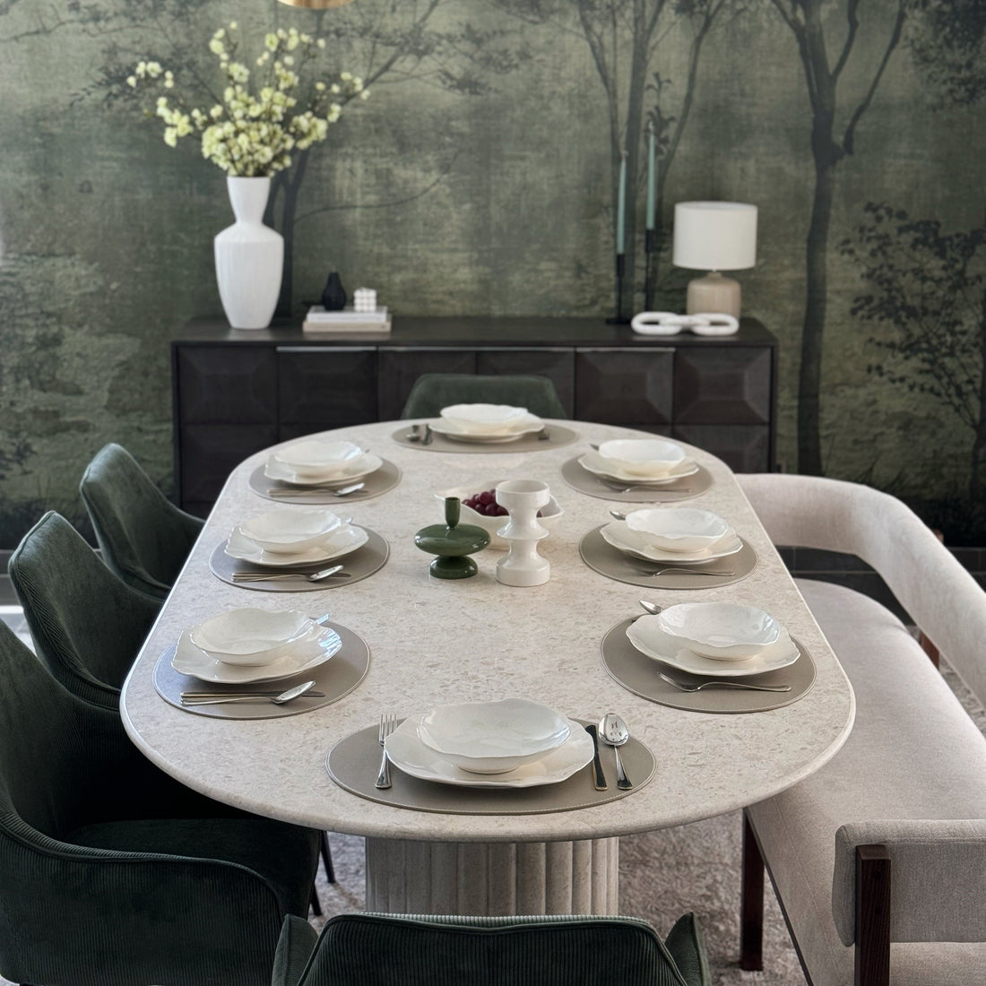 Keti Kerdi Home selection of oval marble dining table, pictured with 8 plates, a classic furniture bench on one long end, 3 green rounded leather seats on the other long side and single rounded green leather seats on each of the narrow ends